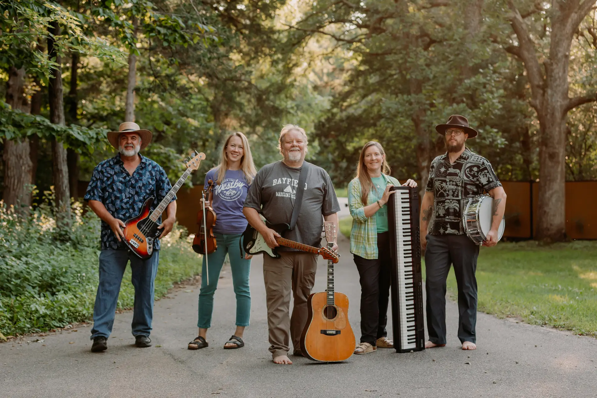 The five members of Mr. Blink standing on a tree-lined driveway holding a bass, fiddle, guitars, keyboard, and snare drum
